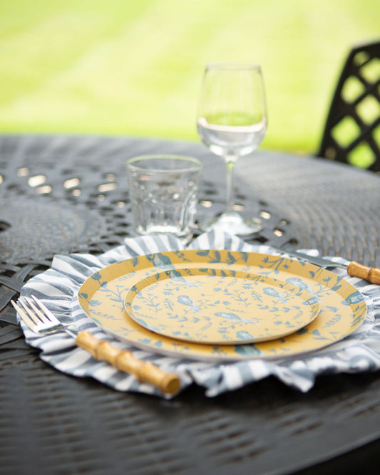Yellow plates with blue patterns on a striped placemat, glassware, and cutlery on a table outdoors.