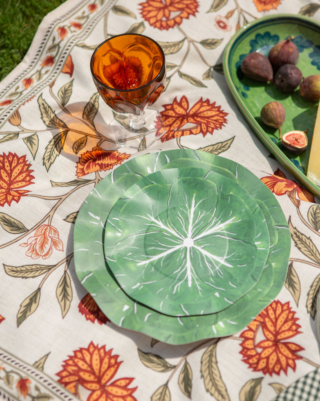 Green leaf-patterned plates on a decorative tablecloth with figs and a glass.