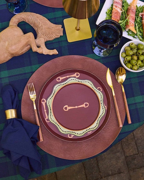 An overhead view of a styled dining table setting featuring a tablecloth in a green and blue plaid pattern, matching brown and burgundy plates with gold accents, gold cutlery, a navy napkin, and a wooden horse centerpiece.