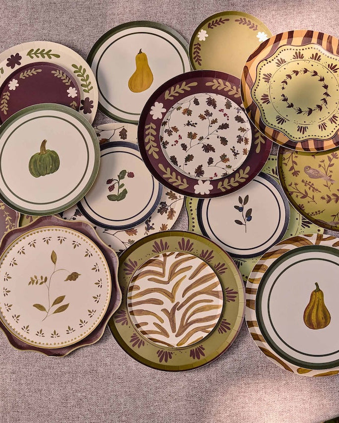 A collection of decorative plates with autumn-themed patterns, including pumpkins, pears, leaves, and floral motifs, arranged on a round gray table.