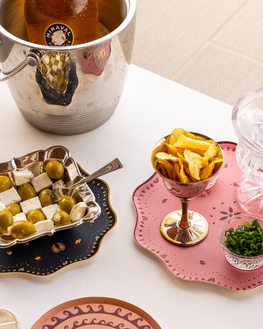 A styled tabletop arrangement of decorative serving dishes and food: a pink scalloped plate holding potato chips in a pedestal bowl, a dark navy plate with a checkerboard olives and feta dish, a peach plate in the foreground, a silver ice bucket, and a glass bowl of greens on a white surface.