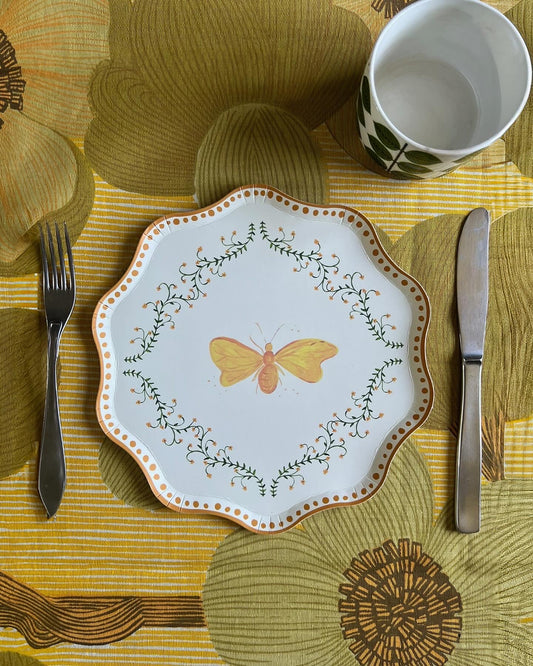 Decorative plate with butterfly design on a patterned tablecloth with cutlery and a cup.