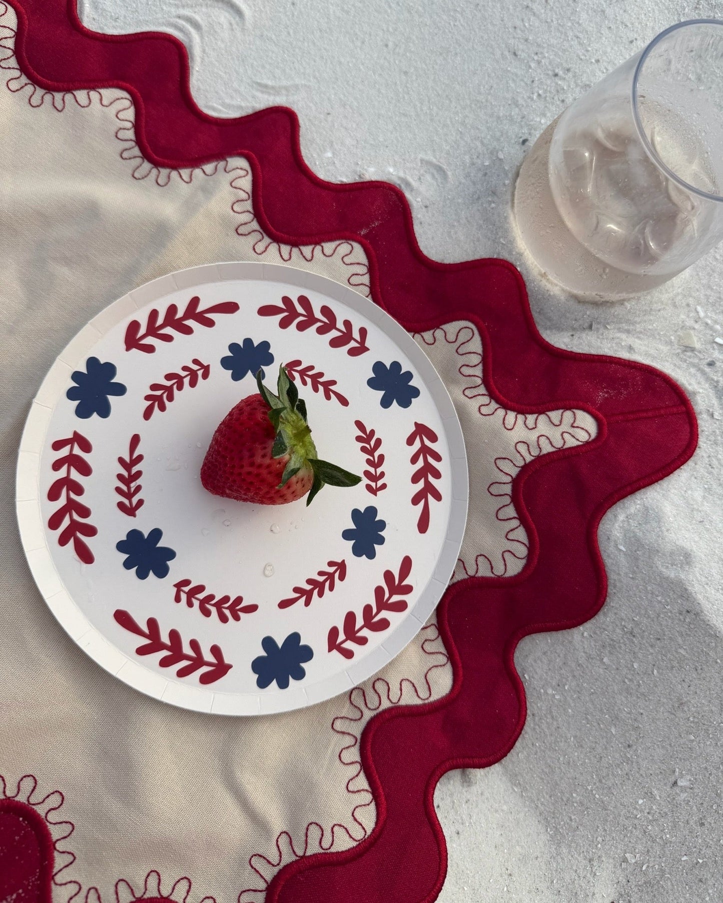 A red and white decorative plate on a cream and red placemat in the sand