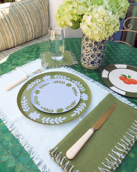 Table setting with blue and green plates, napkins, and a vase of light green flowers on a green table.