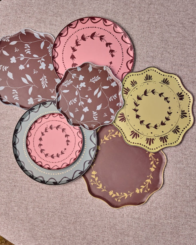 Decorative plates some with scalloped edges are arranged in a cluster on a round table. The plates feature various patterns and colors including pink, brown, yellow, and gray, with some showing floral motifs and others displaying geometric or lace-like borders.