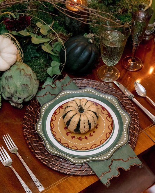 A styled autumn table setting featuring a decorative place setting with a small striped pumpkin centerpiece on a patterned plate, surrounded by a wicker charger, green napkin, cutlery, and a lush fall floral and greenery centerpiece with pumpkins and candles on a wooden table.