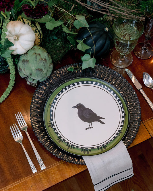 A styled table setting featuring a decorative dinner plate with a black raven illustration, layered over a dark woven charger plate, with silver forks, a napkin, and autumnal floral decor in the background.