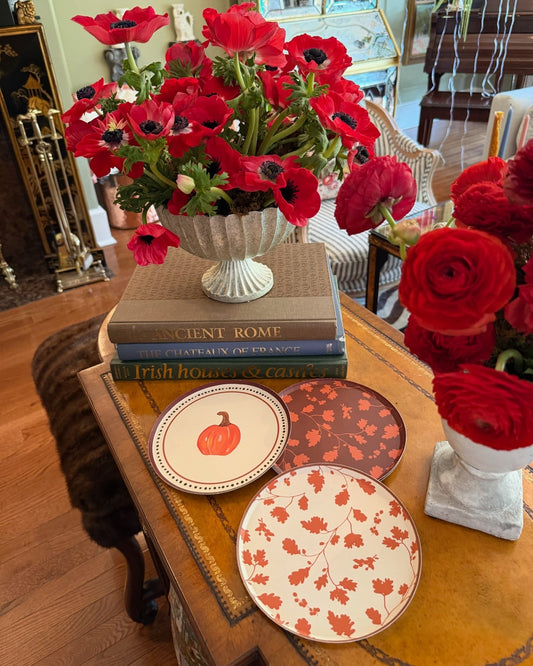 Three decorative plates with autumn-themed patterns (pumpkin, leaves, and maple leaves) arranged on a wooden table next to a white vase of red poppies in a home interior setting.