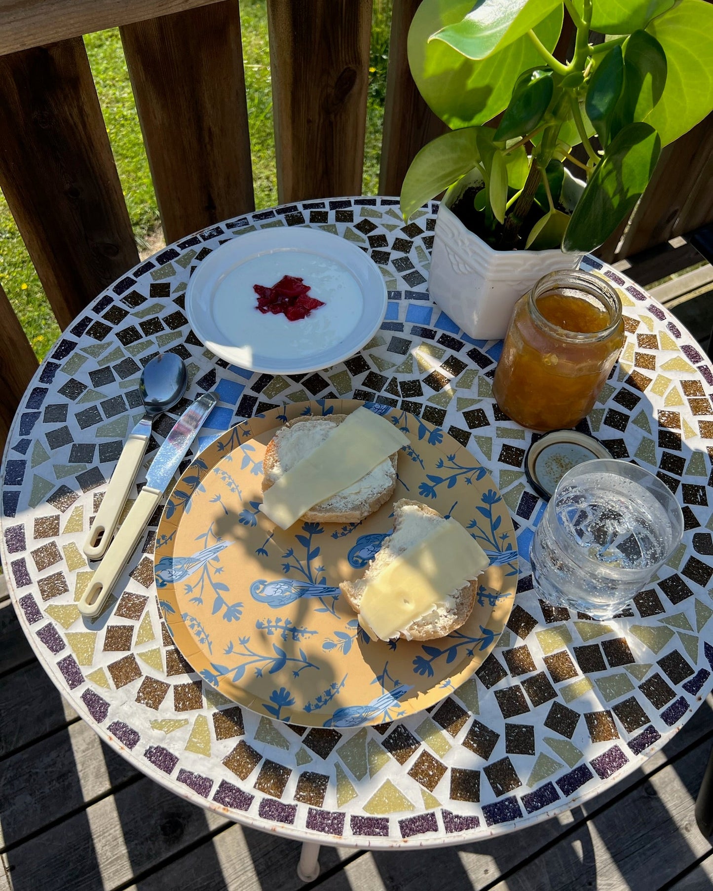 Yellow plate with blue birds on a Mosaic table with bread, and a jar of honey on a wooden deck.