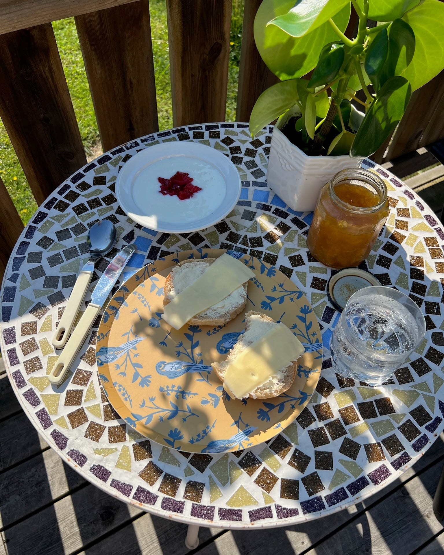 Yellow plate with blue birds on a Mosaic table with bread, and a jar of honey on a wooden deck.