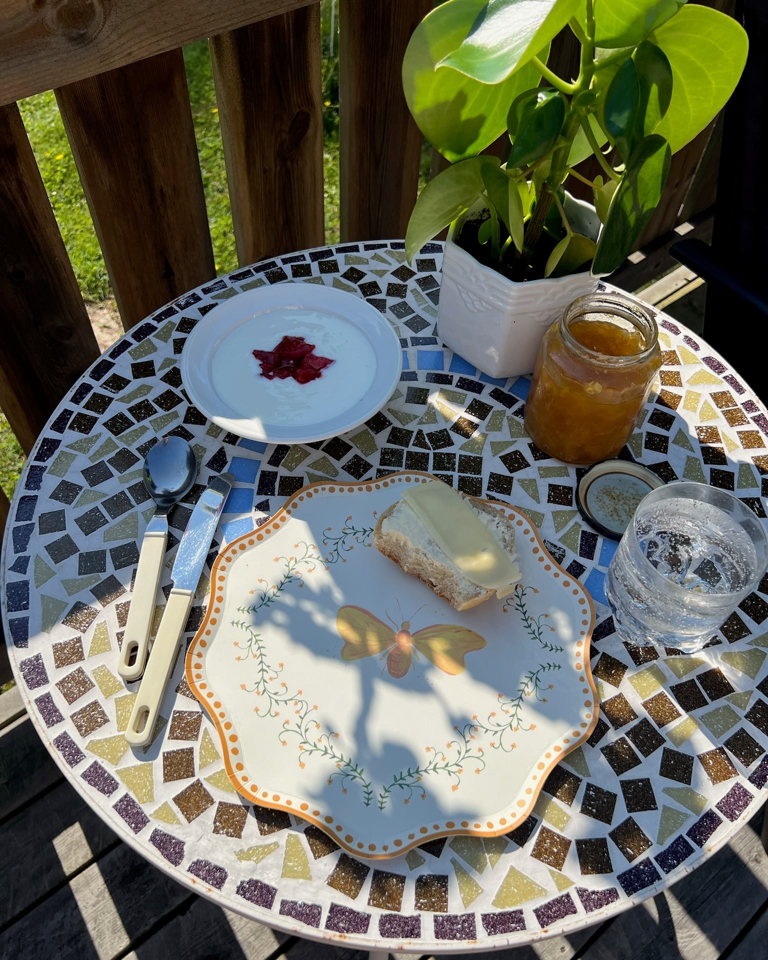Mosaic table with plates, cutlery, and a plant on a wooden deck.