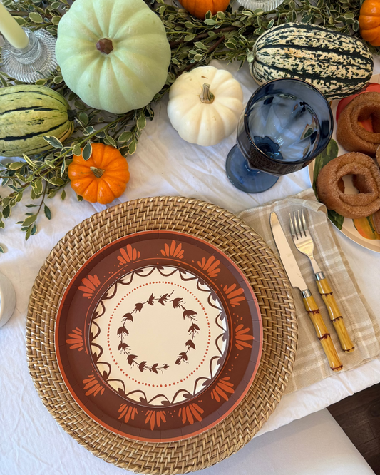 A styled autumn table setting featuring a round woven straw charger plate under a terracotta and cream patterned plate. The setting includes a blue tinted glass, a silver and gold knife, a checkered napkin, and fall decorations like mini pumpkins, a striped gourd, and greenery on a white tablecloth.