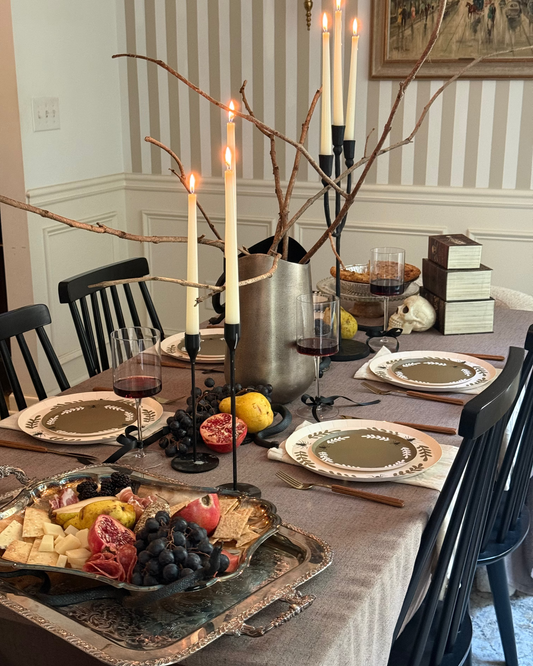 A styled dining table in a home setting, featuring a beige tablecloth, place settings with gold-rimmed plates, a charcuterie board with cheese, grapes, and cured meat, a silver bucket centerpiece with bare branches and lit taper candles, and a striped wall in the background.