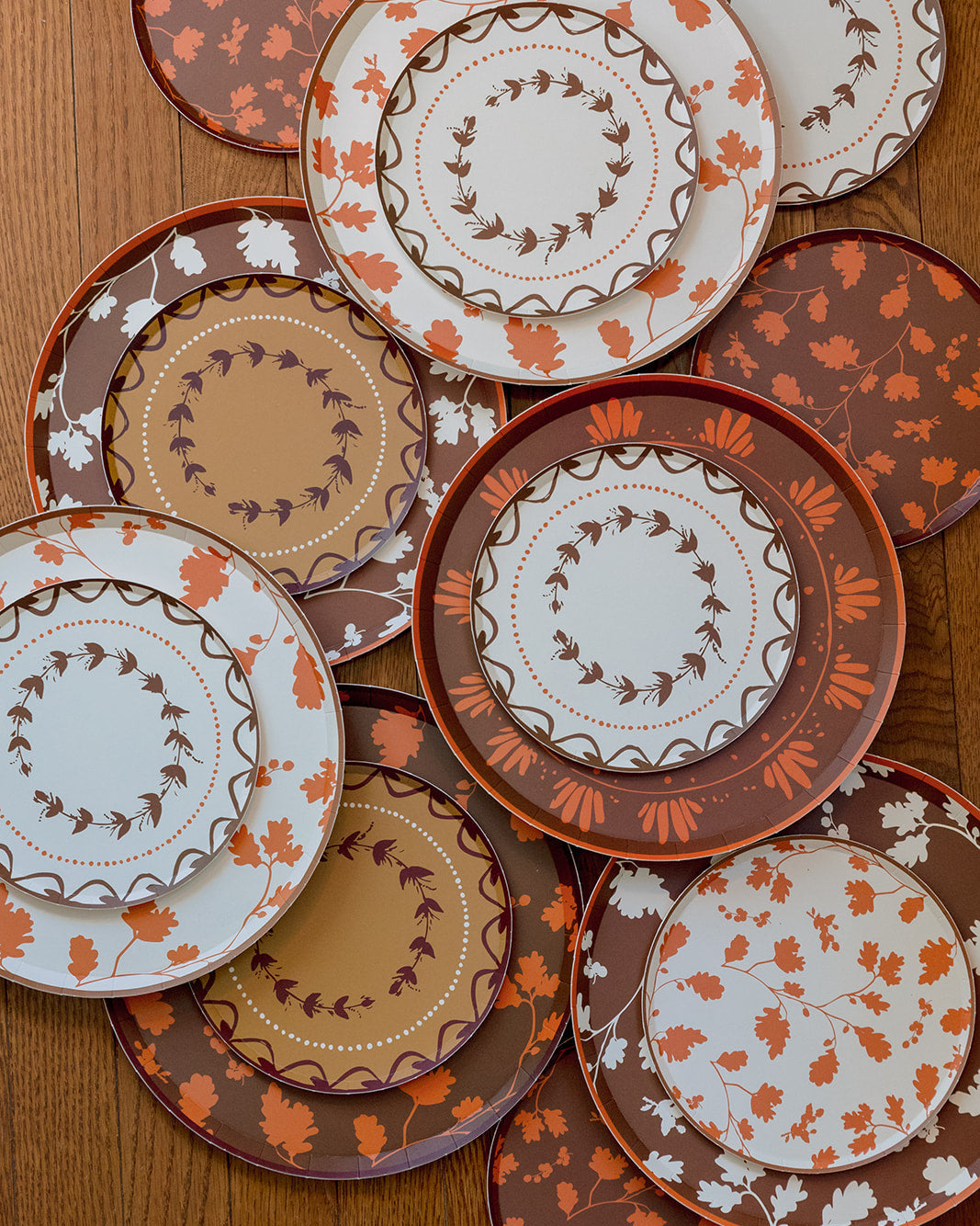 A collection of plates with autumn-themed patterns, including orange and brown leaves, floral motifs, and wreaths on a white background, arranged on a wooden table.