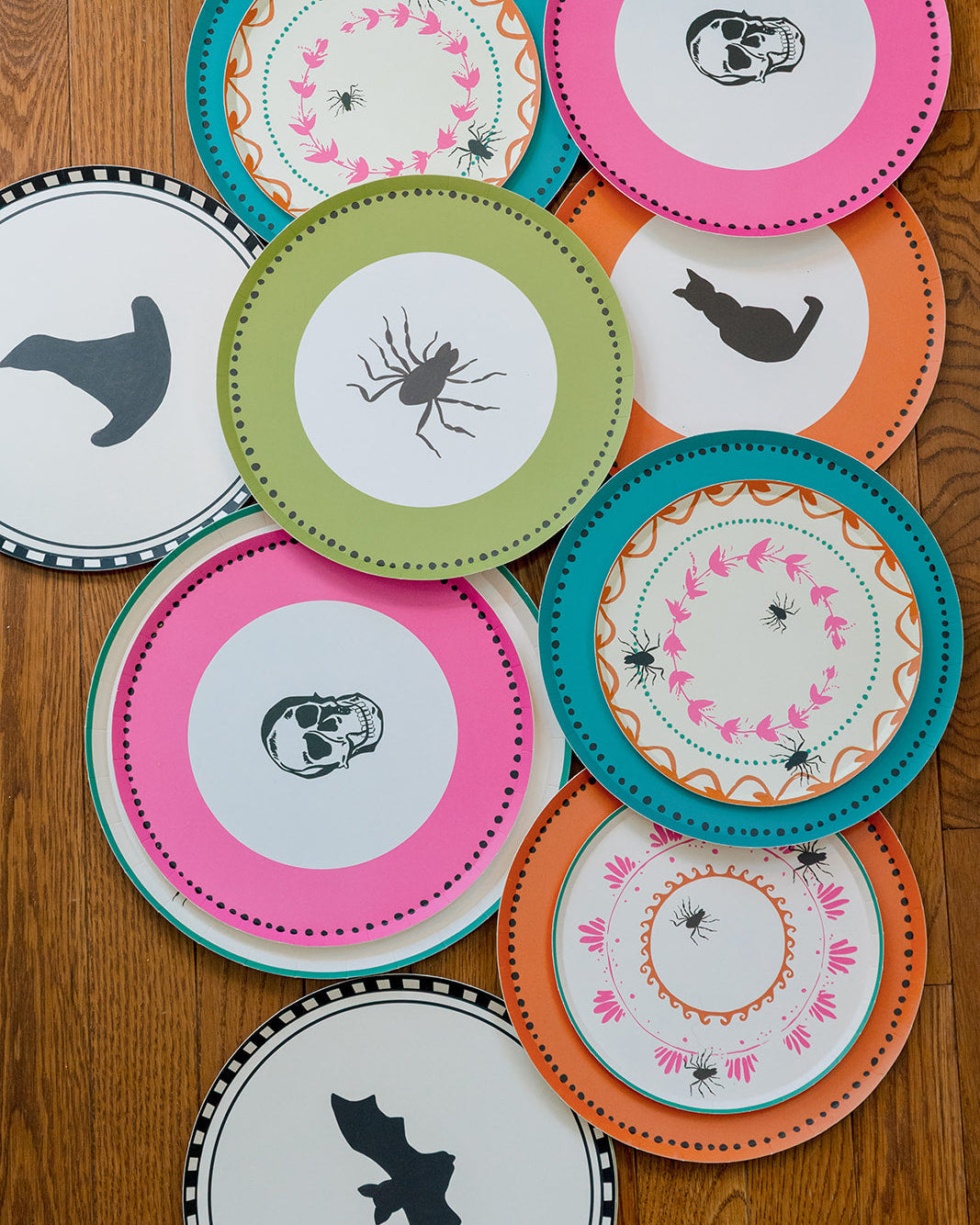 A collection of colorful Halloween-themed paper plates arranged on a wooden tabletop. The plates feature various spooky designs including skulls, bats, spiders, and a witch's hat, with vibrant pink, teal, orange, green, and white colors.