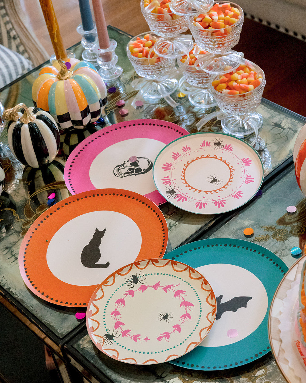 A styled tabletop display of Halloween-themed paper plates in orange, pink, teal, and white, decorated with black cat silhouettes, bats, spiders, and floral patterns. The scene includes candy corn in glass bowls, striped pumpkins, and candles on a glass table.