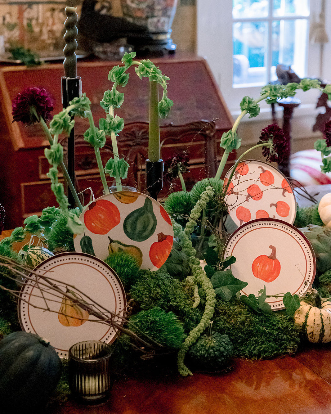 A styled autumn centerpiece on a wooden table, featuring decorative paper plates with orange and green pumpkin and pear illustrations, green textured stems, pinecones, moss, and candles in a rustic indoor setting.