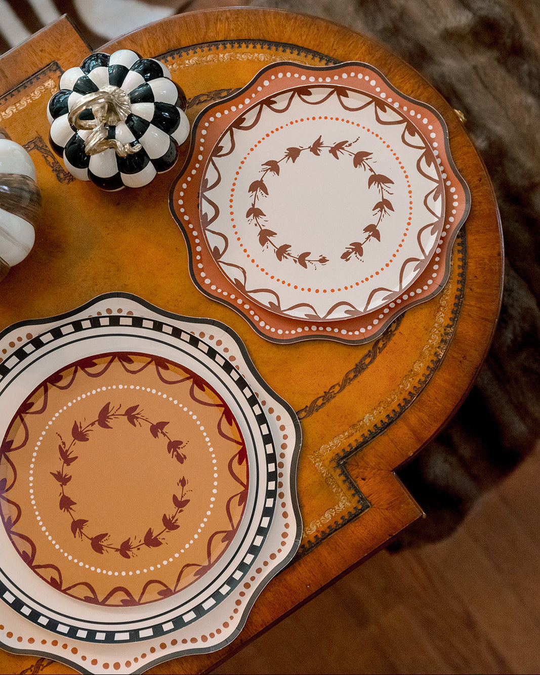 Two scalloped plates with autumn-themed orange, brown, and cream patterns, styled on a wooden side table next to a black and white checkered pumpkin.