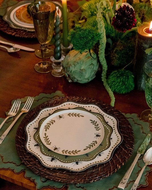 A styled table setting with layered plates featuring a green leaf border, a dark brown woven charger, and green placemats on a wooden table, accented by lit candles and a green floral centerpiece.