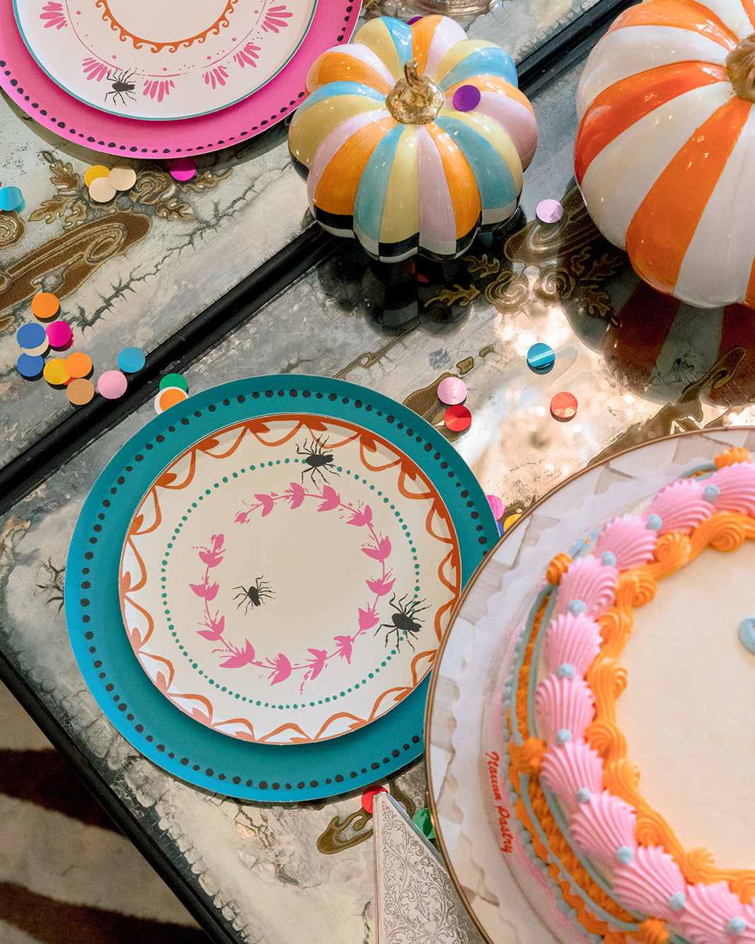 A styled tabletop scene with decorative Halloween-themed plates, a striped pumpkin ornament, confetti, and part of a decorated cake on a reflective mirror surface.