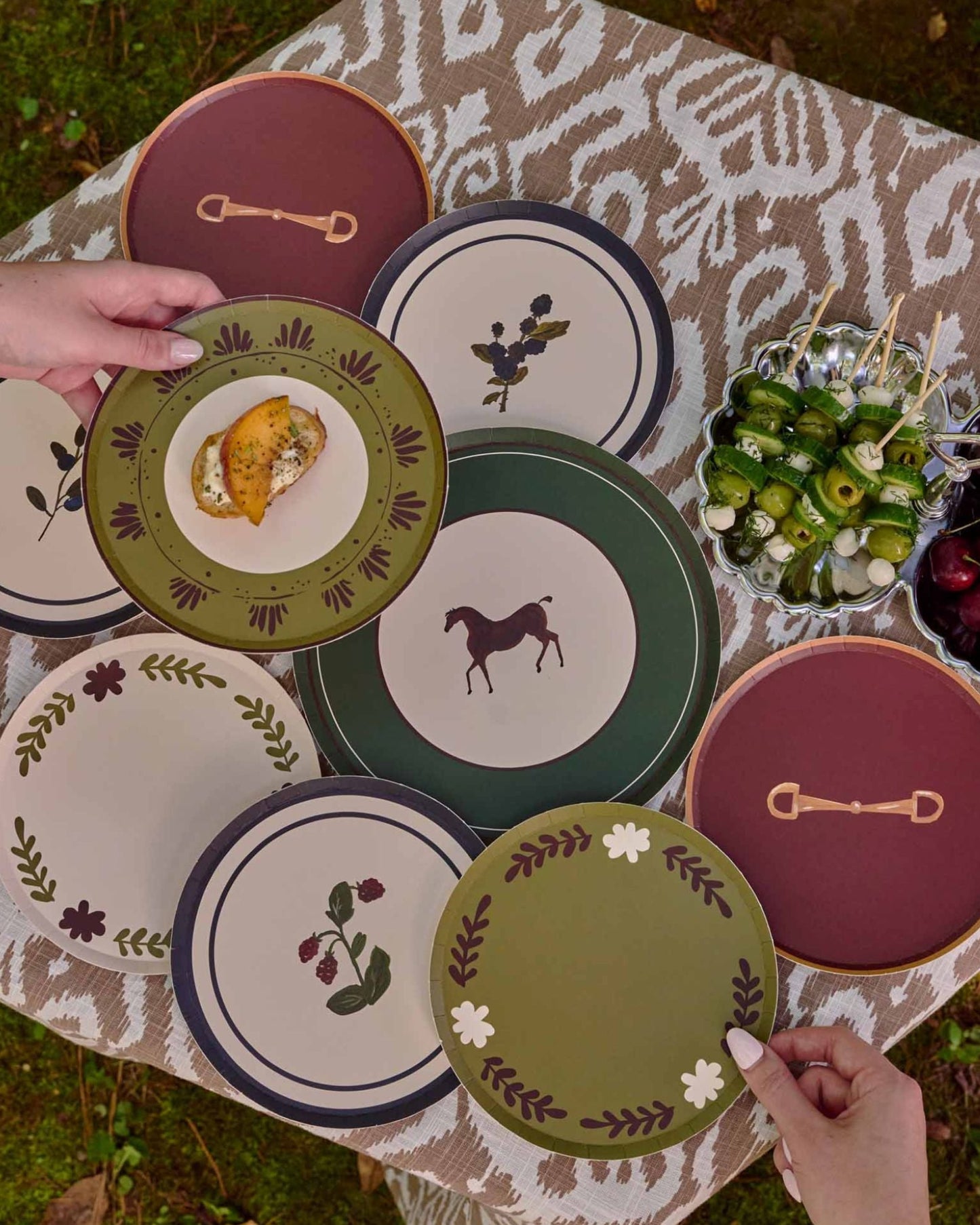 A styled outdoor dining scene showing a collection of decorative plates with horse and botanical designs, arranged on a patterned tablecloth next to a wicker chair on grass.