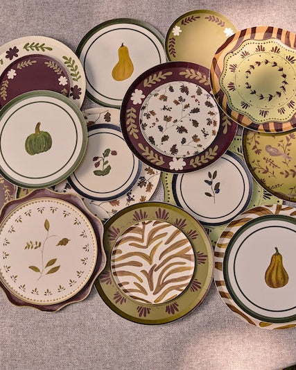 A collection of decorative plates with autumn-themed patterns, including pumpkins, gourds, leaves, and stripes, arranged on a light gray fabric-covered round table.