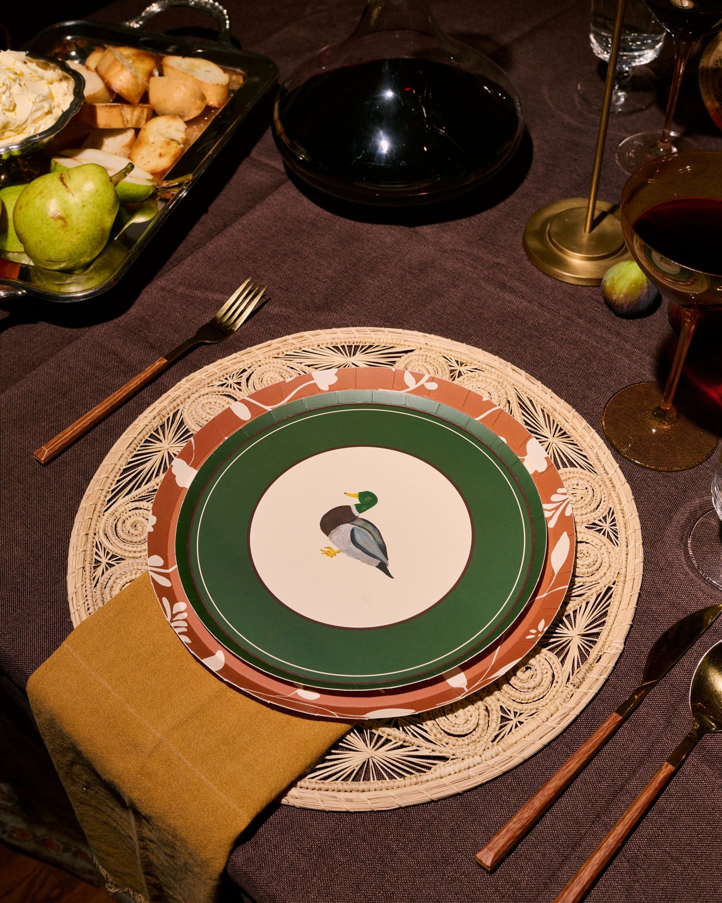 A styled dinner table setting featuring a place setting with a green and white plate printed with a mallard duck, layered over a brown and beige patterned charger plate and a woven placemat. A folded mustard yellow napkin, wooden-handled cutlery, and a dark brown tablecloth complete the rustic table arrangement.