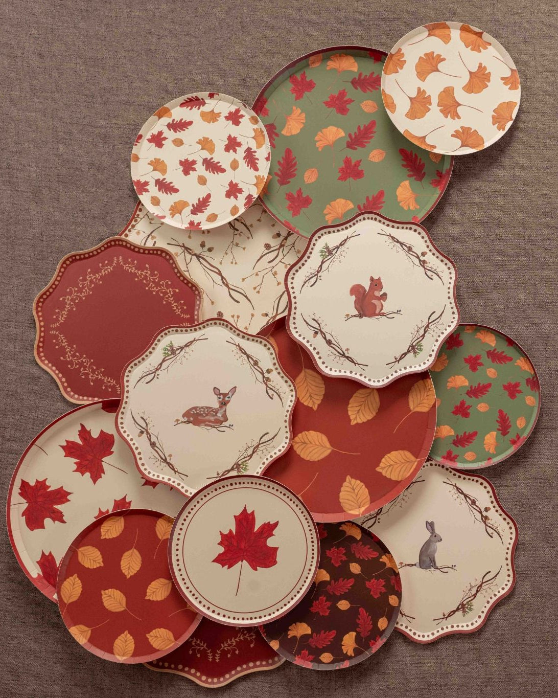 Overhead arrangement of autumn-themed decorative paper plates in cream, rust, burgundy, and sage green, featuring colorful fall leaves, maple leaf prints, and woodland animal illustrations of a deer, squirrel, and rabbit on a textured brown backdrop.