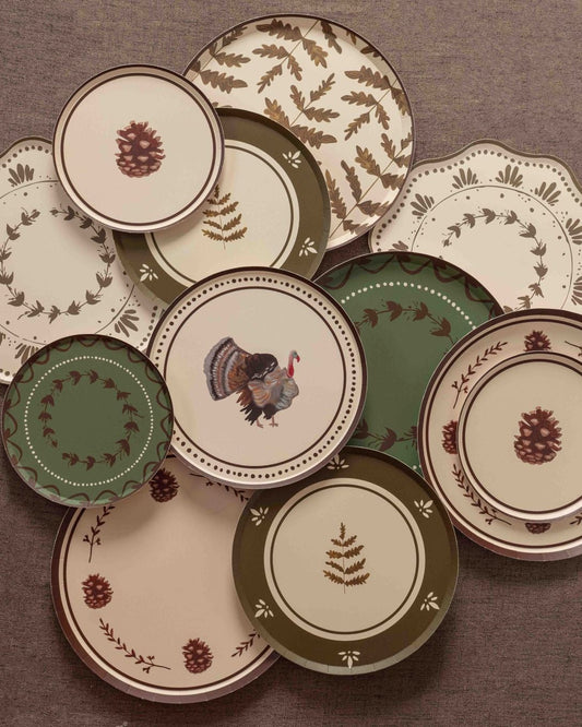 Overhead arrangement of fall-themed paper plates in cream, olive green, and burgundy featuring pinecone illustrations, botanical sprigs, fern patterns, floral wreaths, and a turkey motif on a textured brown backdrop.