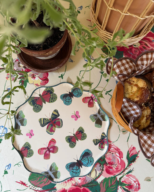 A scalloped plate with a colorful butterfly pattern, styled on a floral tablecloth alongside a potted plant, a wicker basket, and a basket of pastries.