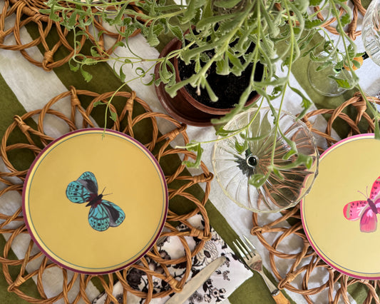 A styled tabletop setting featuring two round plates with butterfly illustrations, woven round placemats, a striped tablecloth, and green foliage.