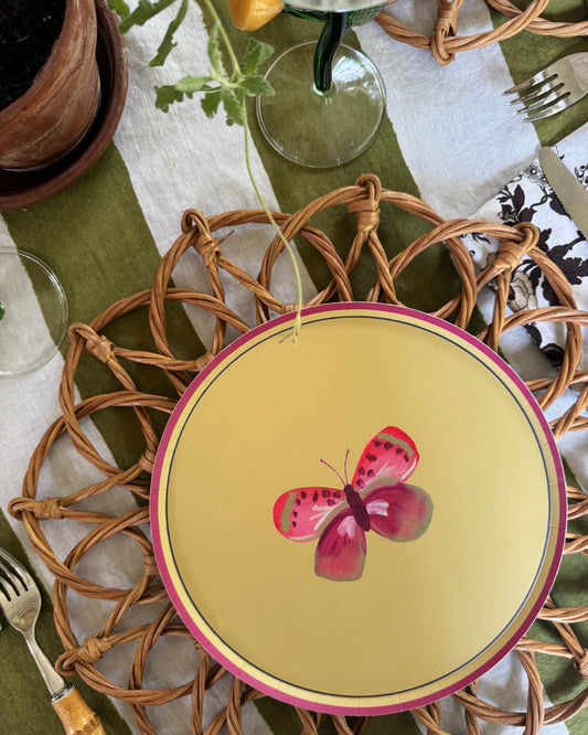 A round green plate with a pink butterfly illustration and pink rim, styled on a woven wicker placemat over a green and white tablecloth with cutlery and glassware.