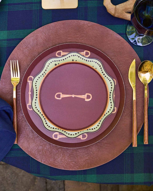 A styled table setting featuring a rustic place setting with a navy blue napkin, gold-toned cutlery, and a layered plate set in terracotta, rust, and olive green on a green plaid tablecloth.