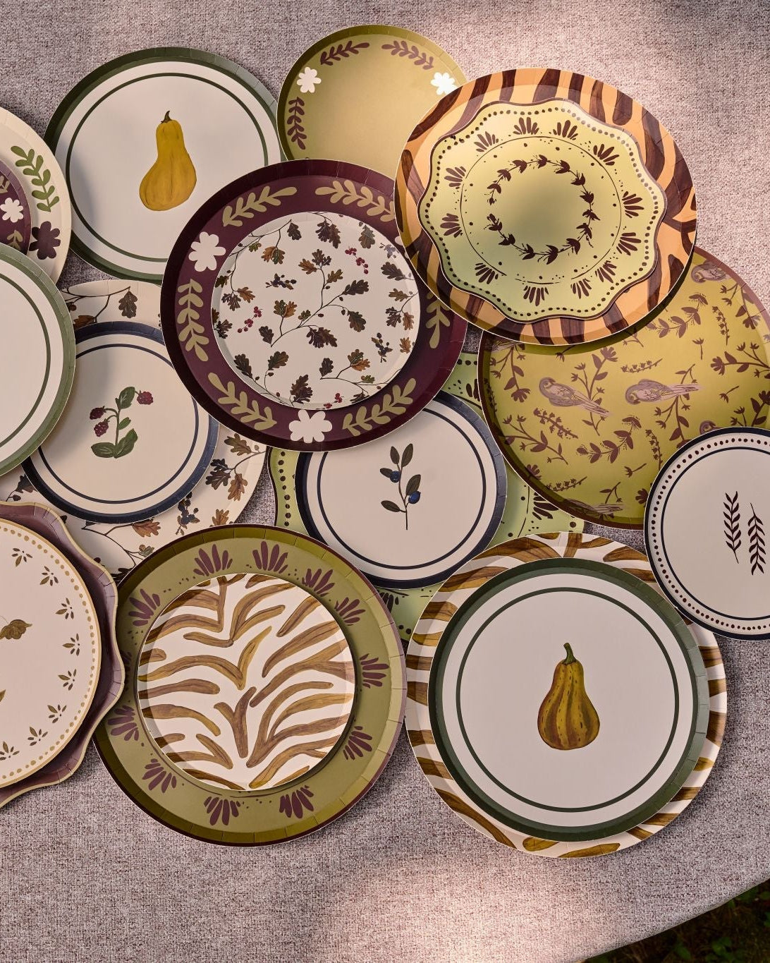A collection of plates with various patterns, including fruit motifs, stripes, and floral designs, arranged on a round gray stone patio outdoors.
