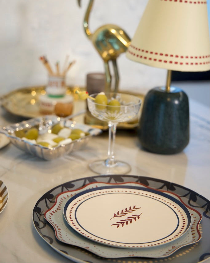 A styled tabletop setting featuring a decorative dinner plate with a red leaf motif in the center. cream-colored table lamp with red dotted trim and a dark green base stands next to a gold flamingo figurine and a glass coupe filled with olives.
