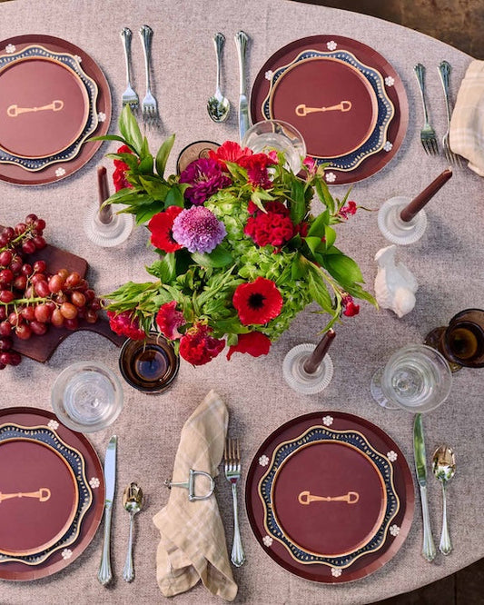 An overhead view of a beautifully set round dining table with 4 place settings, silver cutlery, folded cream napkins, clear drinking glasses, a central floral centerpiece, and a small wooden board of red grapes on a textured beige tablecloth.