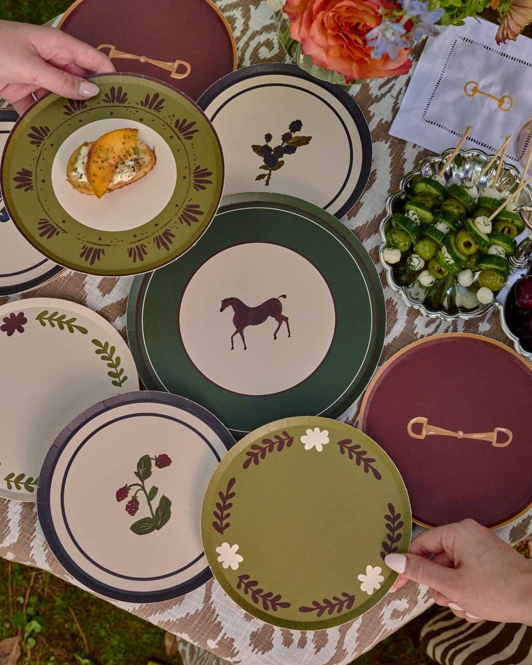 A top-down outdoor view of a table covered with multiple decorative plates in green, cream, and burgundy, with hands holding plates. Bowls of fruit and a wicker basket are visible on a grassy lawn.