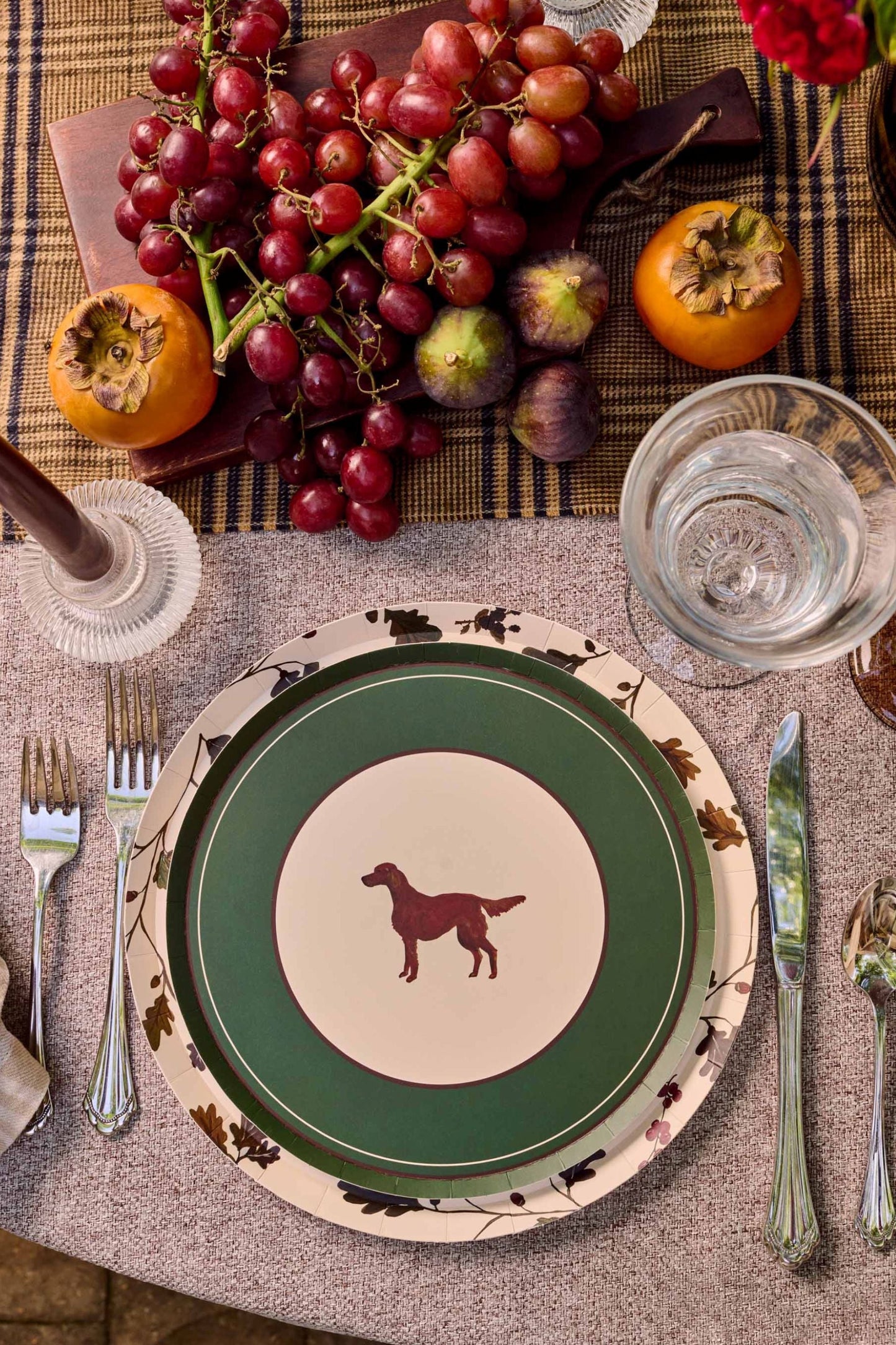An overhead styled table setting featuring a round plate with a green and cream design and a dog silhouette in the center, surrounded by silver cutlery, a beige napkin, and autumn table decor including persimmon, grapes, and a candle.