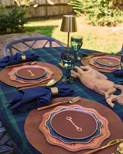 An outdoor table setting with a green and blue plaid tablecloth, layered brown and copper plates, navy napkins with gold rings, gold cutlery, and a rustic deer head centerpiece.