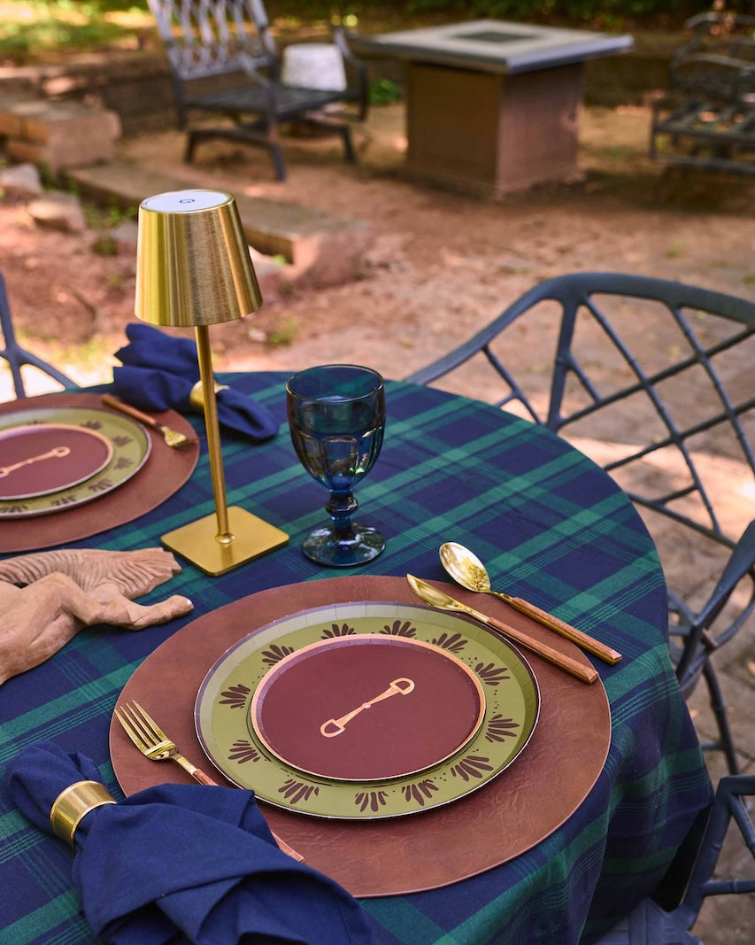 Outdoor dining table set with a green and teal plaid tablecloth, matching napkins, gold-toned flatware, and decorative plates on dark metal patio chairs.