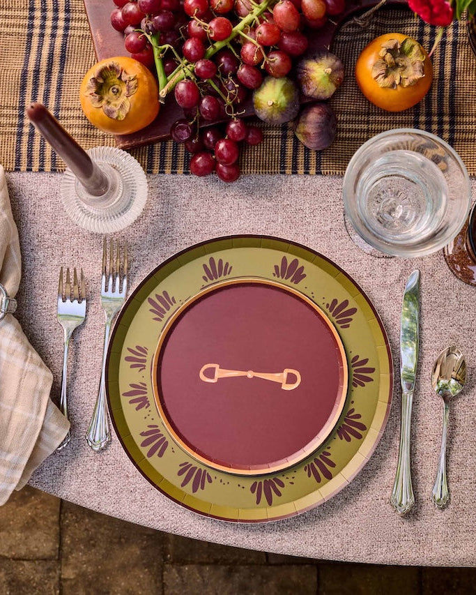A styled outdoor table setting with a place setting featuring a deep red plate with a gold key motif, surrounded by a green and gold patterned charger plate. The table is decorated with a floral centerpiece, red candles, glasses, and fresh fruit.