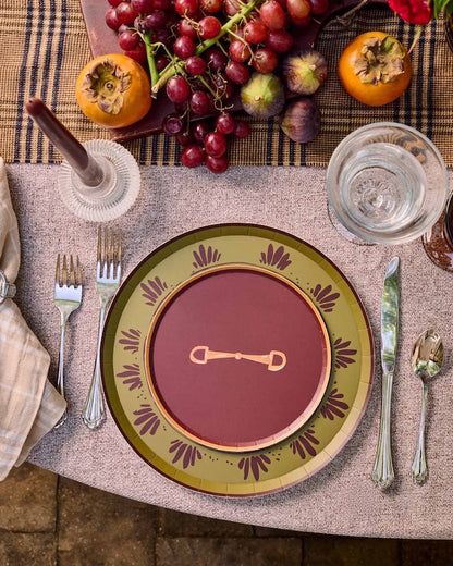 A styled outdoor table setting with a place setting featuring a deep red plate with a gold key motif, surrounded by a green and gold patterned charger plate. The table is decorated with a floral centerpiece, red candles, glasses, and fresh fruit.