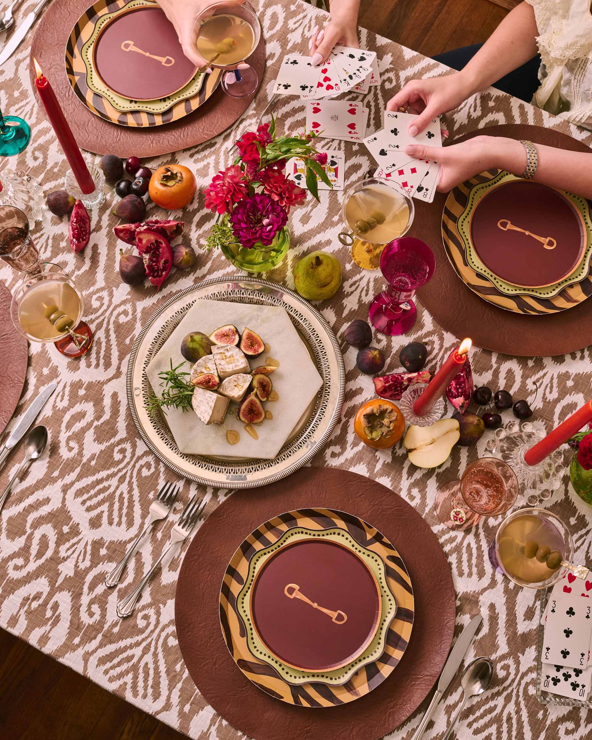 An overhead view of a styled dining table with multiple place settings, patterned tablecloths, candles, fruit, and hands holding playing cards. The tableware features a coordinated color palette of deep burgundy, brown, gold, and black in an ornate pattern.