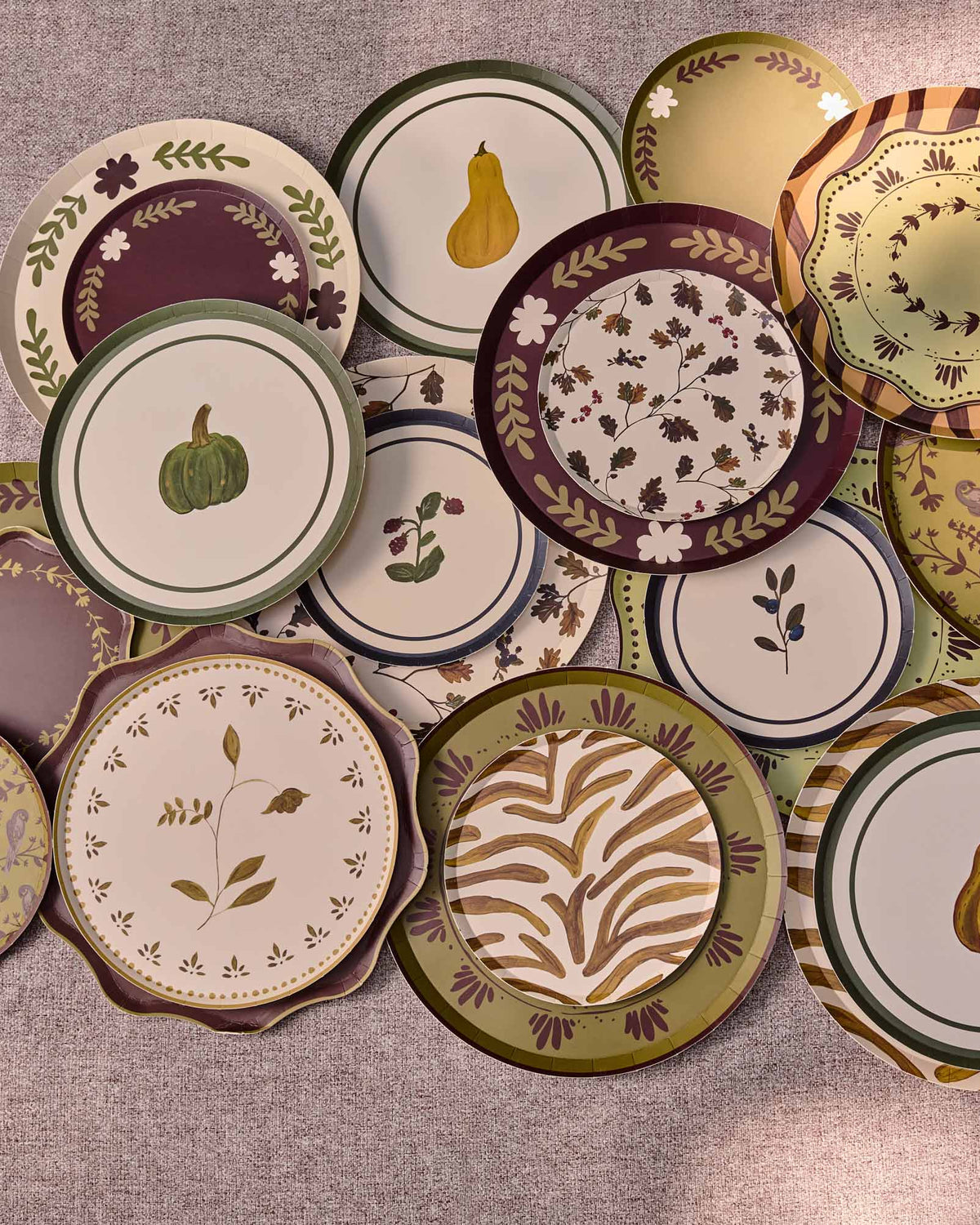 A collection of decorative plates with autumn-themed patterns, including pumpkins, pears, leaves, and floral motifs, arranged on a round gray table.