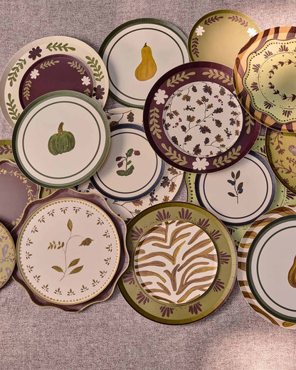 A collection of decorative plates with autumn-themed patterns, including pumpkins, pears, leaves, and floral motifs, arranged on a round gray table.