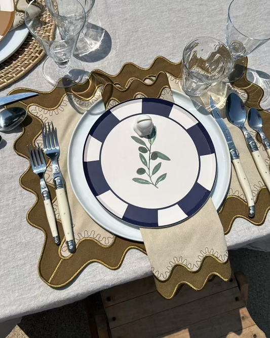Table setting with a decorative plate featuring an olive branch design, surrounded by cutlery and glasses on a textured surface.