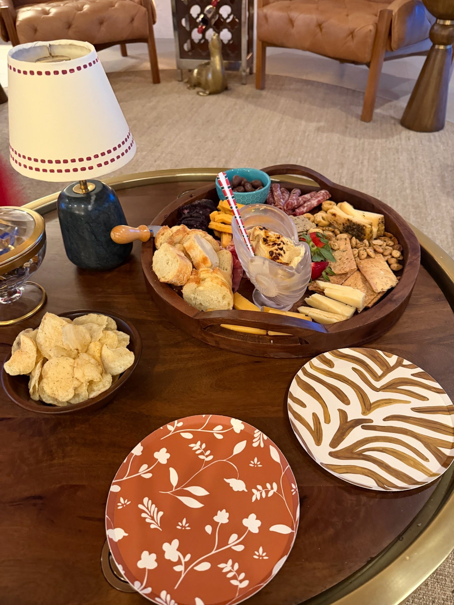 A styled tabletop serving scene with a large wooden charcuterie board filled with breads, cheeses, crackers, and a drink, alongside smaller bowls of chips and patterned plates on a round wooden table in an indoor lounge setting.