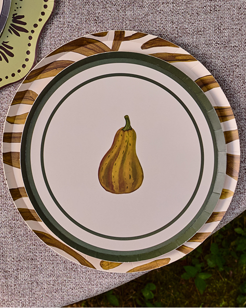 A round paper plate with a central painted squash illustration, surrounded by concentric green rings and a striped outer border, placed on a textured gray surface. Part of another decorative plate is visible at the top left.