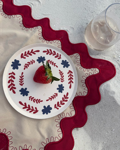 A red and white decorative plate on a cream and red placemat in the sand