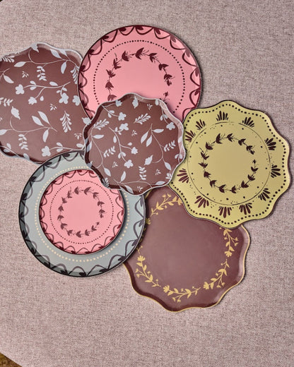 Decorative plates some with scalloped edges are arranged in a cluster on a round table. The plates feature various patterns and colors including pink, brown, yellow, and gray, with some showing floral motifs and others displaying geometric or lace-like borders.